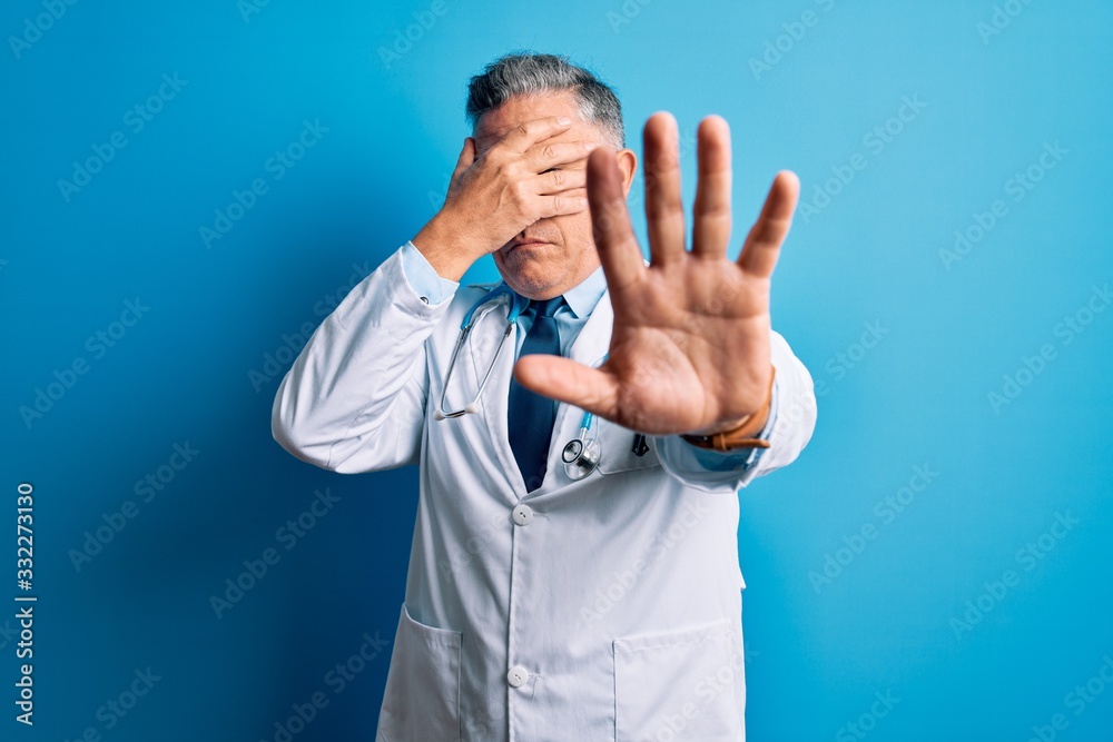 Middle age handsome grey-haired doctor man wearing coat and blue stethoscope covering eyes with hands and doing stop gesture with sad and fear expression. Embarrassed and negative concept. Middle age handsome grey-haired doctor man wearing coat and blue stethoscope covering eyes with hands and doing stop gesture with sad and fear expression. Embarrassed and negative concept.
