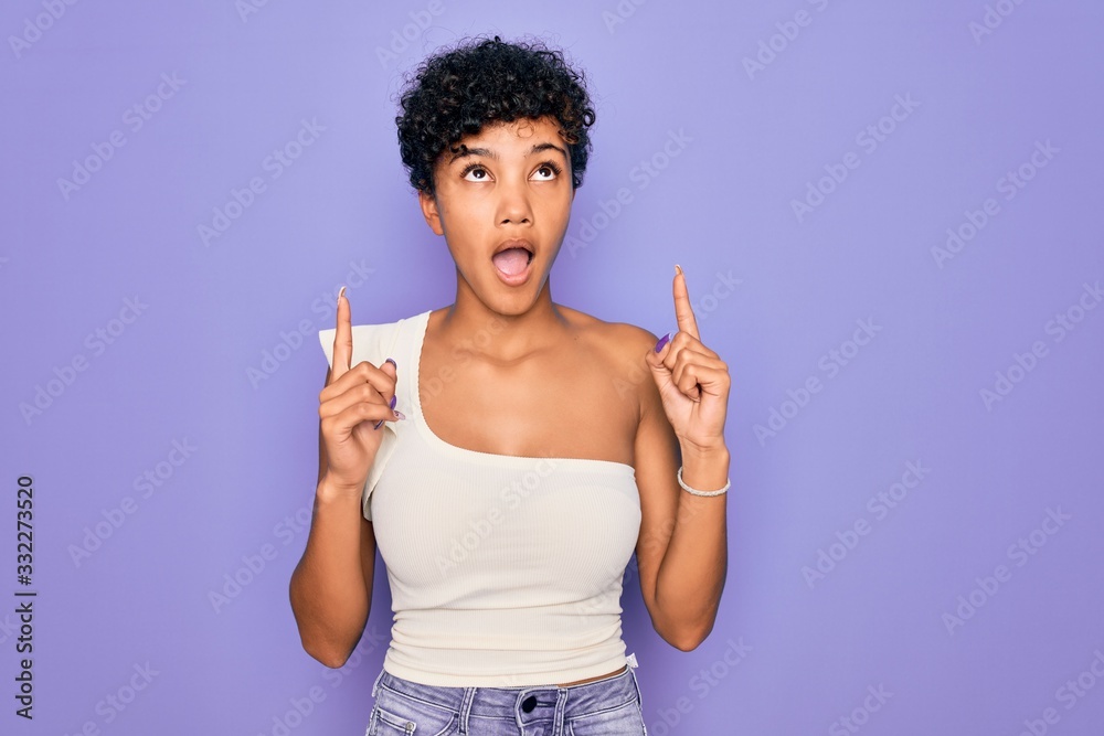 Young beautiful african american afro woman wearing casual t-shirt over purple background amazed and surprised looking up and pointing with fingers and raised arms. Young beautiful african american afro woman wearing casual t-shirt over purple background amazed and surprised looking up and pointing with fingers and raised arms.