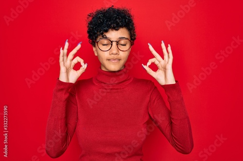 Young beautiful african american afro woman wearing turtleneck sweater and glasses relaxed and smiling with eyes closed doing meditation gesture with fingers. Yoga concept.