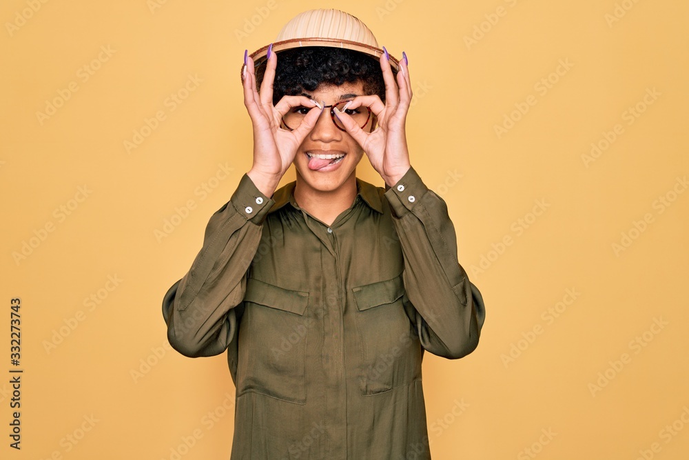 Young beautiful african american tourist woman wearing glasses and explorer hat doing ok gesture like binoculars sticking tongue out, eyes looking through fingers. Crazy expression. Young beautiful african american tourist woman wearing glasses and explorer hat doing ok gesture like binoculars sticking tongue out, eyes looking through fingers. Crazy expression.