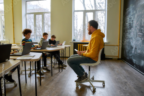 Modern lesson. Portrait of young male teacher communicating with his pupils, while sitting with tablet pc near the blackboard during a lesson in modern smart school