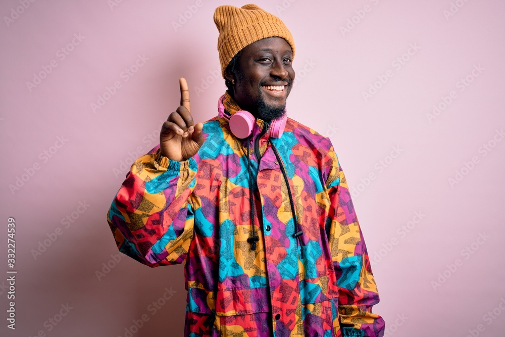 Young handsome african american man wearing colorful coat and cap over pink background showing and pointing up with finger number one while smiling confident and happy.