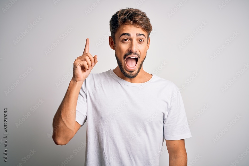 Young handsome man with beard wearing casual t-shirt standing over white background pointing finger up with successful idea. Exited and happy. Number one.