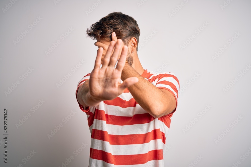 Young handsome man with beard wearing striped t-shirt standing over white background covering eyes with hands and doing stop gesture with sad and fear expression. Embarrassed and negative concept. Young handsome man with beard wearing striped t-shirt standing over white background covering eyes with hands and doing stop gesture with sad and fear expression. Embarrassed and negative concept.