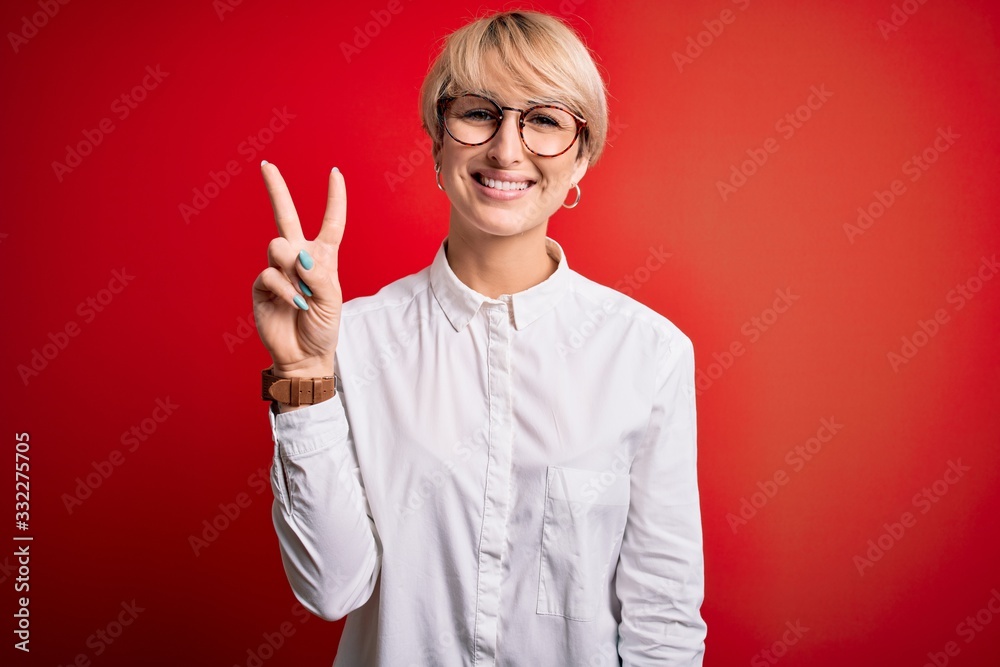 Young blonde business woman with short hair wearing glasses over red background showing and pointing up with fingers number two while smiling confident and happy. Young blonde business woman with short hair wearing glasses over red background showing and pointing up with fingers number two while smiling confident and happy.