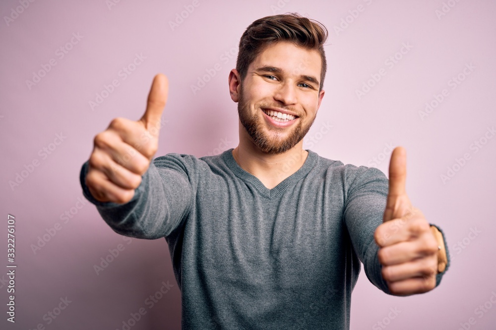 Young handsome blond man with beard and blue eyes wearing casual sweater approving doing positive gesture with hand, thumbs up smiling and happy for success. Winner gesture. Young handsome blond man with beard and blue eyes wearing casual sweater approving doing positive gesture with hand, thumbs up smiling and happy for success. Winner gesture.
