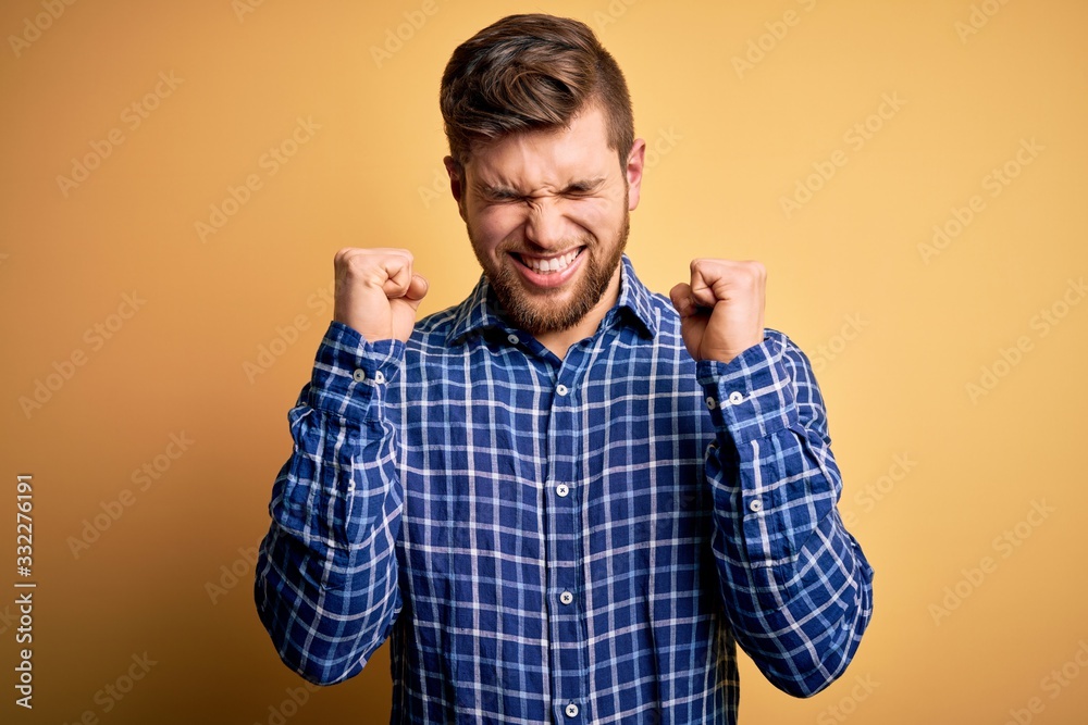 Young blond businessman with beard and blue eyes wearing shirt over yellow background excited for success with arms raised and eyes closed celebrating victory smiling. Winner concept. Young blond businessman with beard and blue eyes wearing shirt over yellow background excited for success with arms raised and eyes closed celebrating victory smiling. Winner concept.