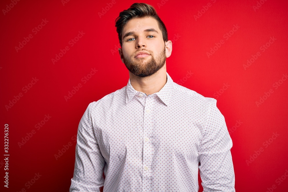 Young blond businessman with beard and blue eyes wearing elegant shirt over red background Relaxed with serious expression on face. Simple and natural looking at the camera. Young blond businessman with beard and blue eyes wearing elegant shirt over red background Relaxed with serious expression on face. Simple and natural looking at the camera.