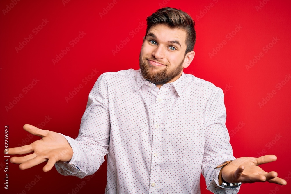 Young blond businessman with beard and blue eyes wearing elegant shirt over red background clueless and confused expression with arms and hands raised. Doubt concept. Young blond businessman with beard and blue eyes wearing elegant shirt over red background clueless and confused expression with arms and hands raised. Doubt concept.