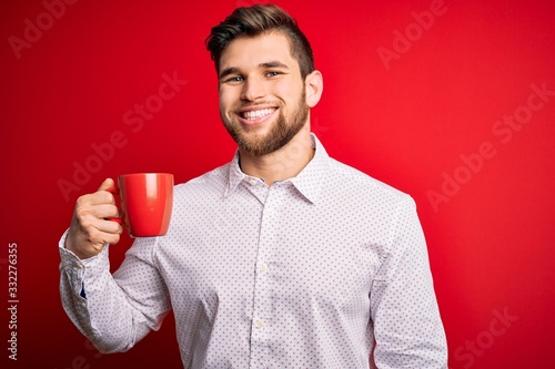 Young blond businessman with beard and blue eyes drinking red cup of coffee with a happy face standing and smiling with a confident smile showing teeth © Krakenimages.com