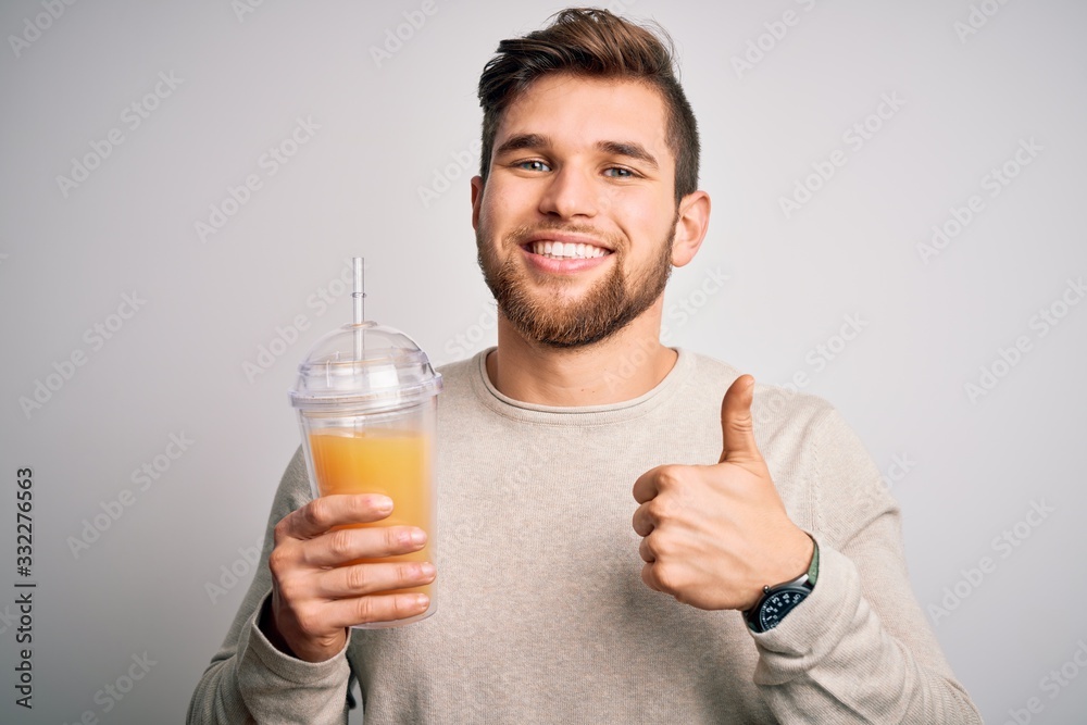 Young blond man with beard and blue eyes drinking healthy orange smoothie happy with big smile doing ok sign, thumb up with fingers, excellent sign Young blond man with beard and blue eyes drinking healthy orange smoothie happy with big smile doing ok sign, thumb up with fingers, excellent sign
