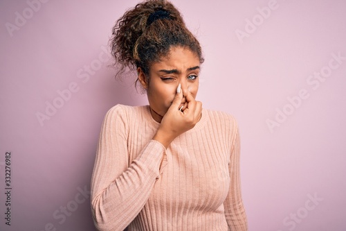 Young beautiful african american girl wearing casual sweater standing over pink background smelling something stinky and disgusting, intolerable smell, holding breath with fingers on nose. Bad smell photo
