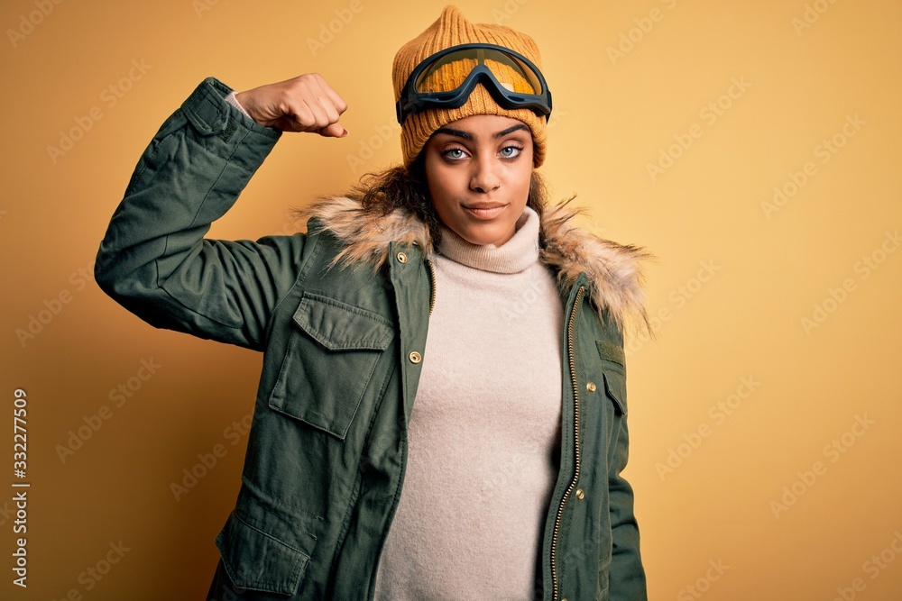 Young african american afro skier girl wearing snow sportswear and ski goggles Strong person showing arm muscle, confident and proud of power Young african american afro skier girl wearing snow sportswear and ski goggles Strong person showing arm muscle, confident and proud of power