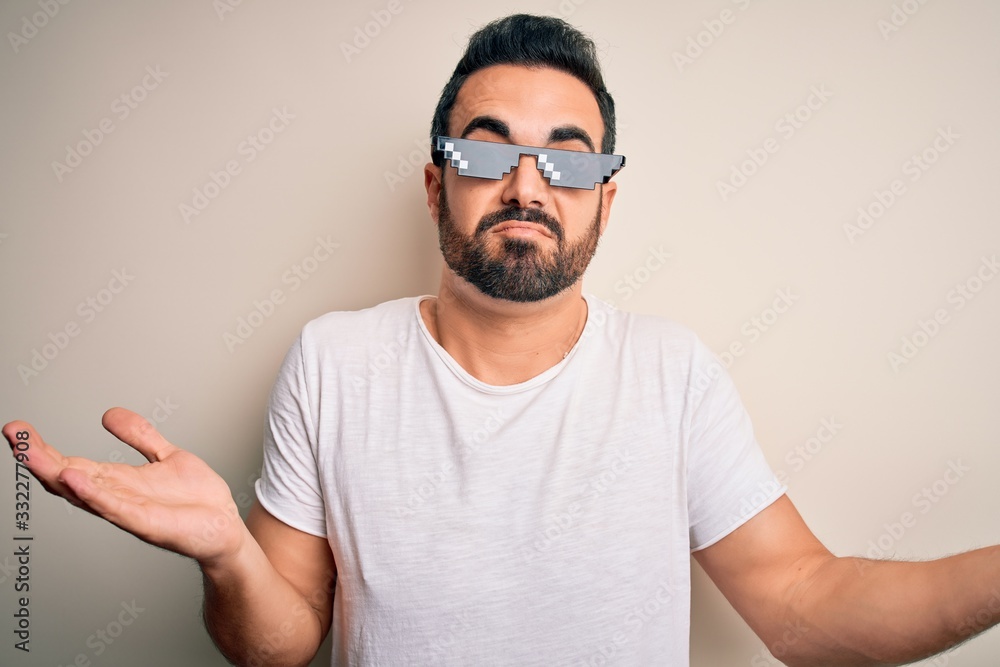 Young handsome man with beard wearing funny thug life sunglasses over white background clueless and confused expression with arms and hands raised. Doubt concept. Young handsome man with beard wearing funny thug life sunglasses over white background clueless and confused expression with arms and hands raised. Doubt concept.