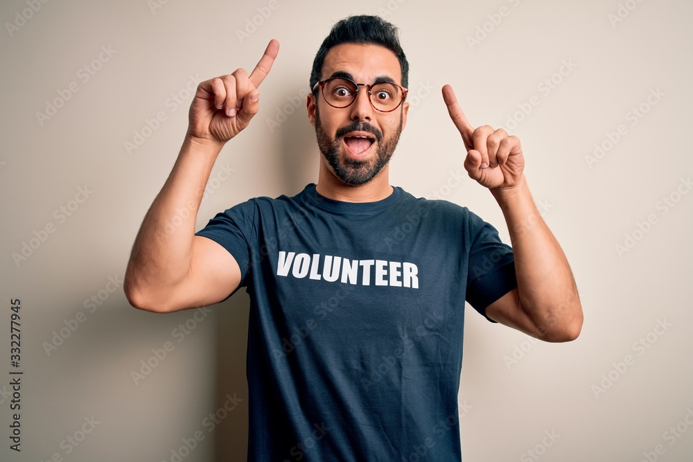 Fototapeta premium Handsome man with beard wearing t-shirt with volunteer message over white background smiling amazed and surprised and pointing up with fingers and raised arms.