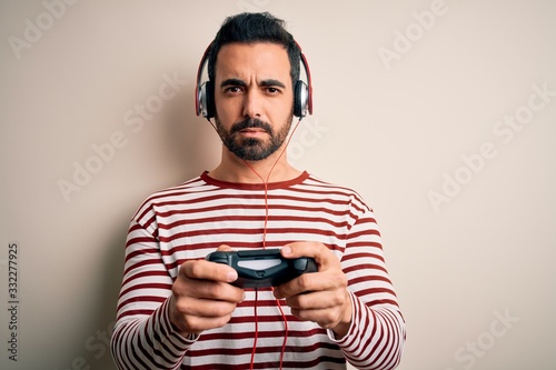 Young handsome gamer man with beard playing video game using joystick and headphones with a confident expression on smart face thinking serious