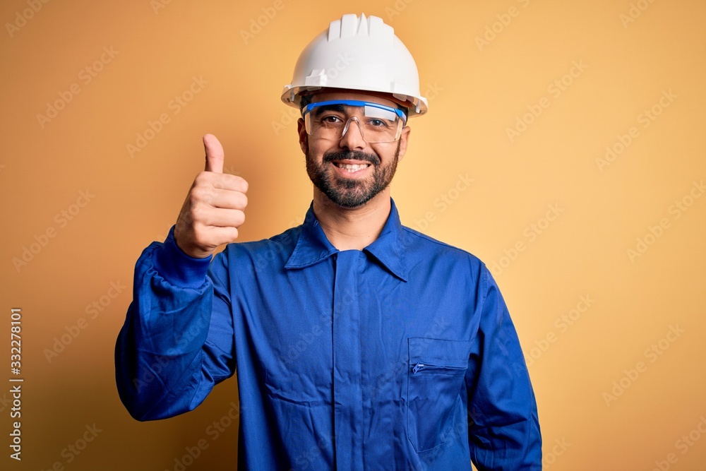 Mechanic man with beard wearing blue uniform and safety glasses over yellow background doing happy thumbs up gesture with hand. Approving expression looking at the camera showing success.