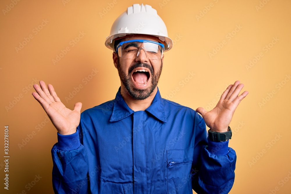 Mechanic man with beard wearing blue uniform and safety glasses over yellow background celebrating mad and crazy for success with arms raised and closed eyes screaming excited. Winner concept