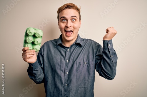 Young handsome redhead man holding box of healthy eggs over isolated white background screaming proud, celebrating victory and success very excited with raised arms