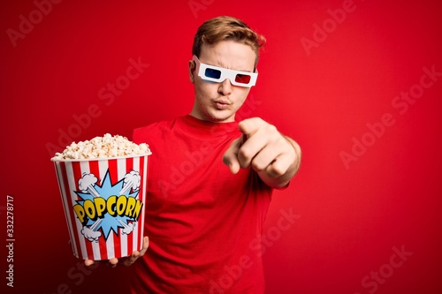 Young handsome redhead man watching 3d glasses eating popcorn snack over red background pointing with finger to the camera and to you, hand sign, positive and confident gesture from the front