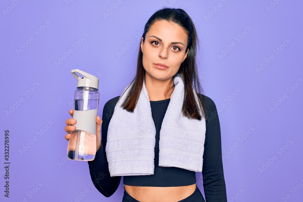 Young beautiful brunette sporty woman using sport towel drinking bottle of water thinking attitude and sober expression looking self confident Young beautiful brunette sporty woman using sport towel drinking bottle of water thinking attitude and sober expression looking self confident