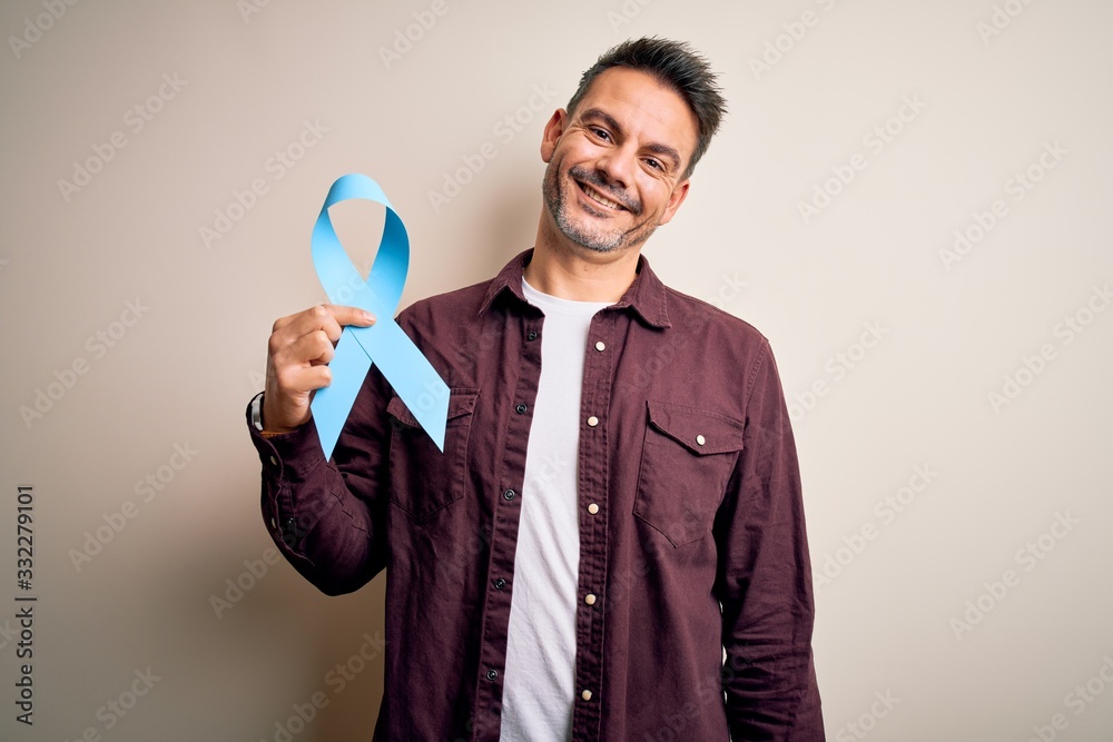 Young handsome man holding blue cancer ribbon over isolated white background with a happy face standing and smiling with a confident smile showing teeth Young handsome man holding blue cancer ribbon over isolated white background with a happy face standing and smiling with a confident smile showing teeth