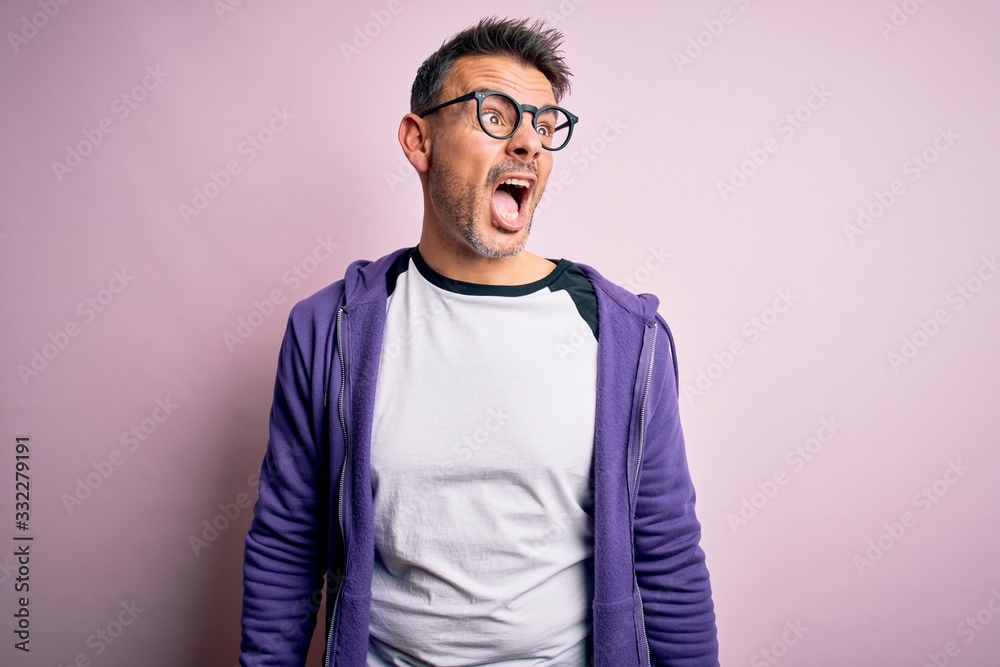 Young handsome man wearing purple sweatshirt and glasses standing over pink background angry and mad screaming frustrated and furious, shouting with anger. Rage and aggressive concept. Young handsome man wearing purple sweatshirt and glasses standing over pink background angry and mad screaming frustrated and furious, shouting with anger. Rage and aggressive concept.