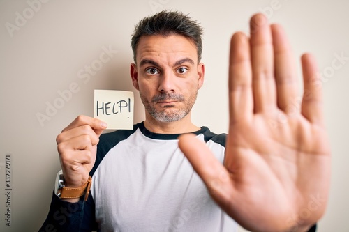 Young handsome man stressed holding reminder paper with help message with open hand doing stop sign with serious and confident expression, defense gesture © Krakenimages.com