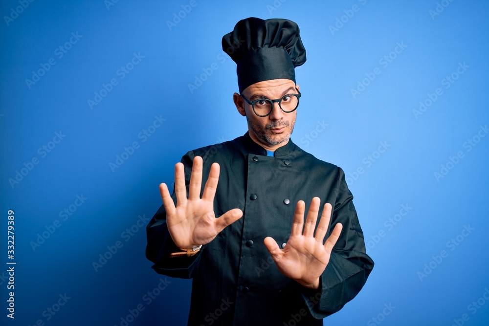 Young handsome chef man wearing cooker uniform and hat over isolated blue background Moving away hands palms showing refusal and denial with afraid and disgusting expression. Stop and forbidden.