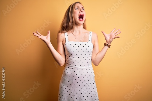 Young beautiful blonde woman on vacation wearing summer dress over yellow background crazy and mad shouting and yelling with aggressive expression and arms raised. Frustration concept.