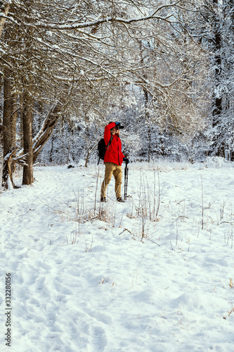Male photographer in red jacket taking pictures of winter landscape in snowy forest.