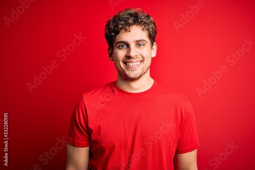 Young blond handsome man with curly hair wearing casual t-shirt over red background with a happy and cool smile on face. Lucky person.