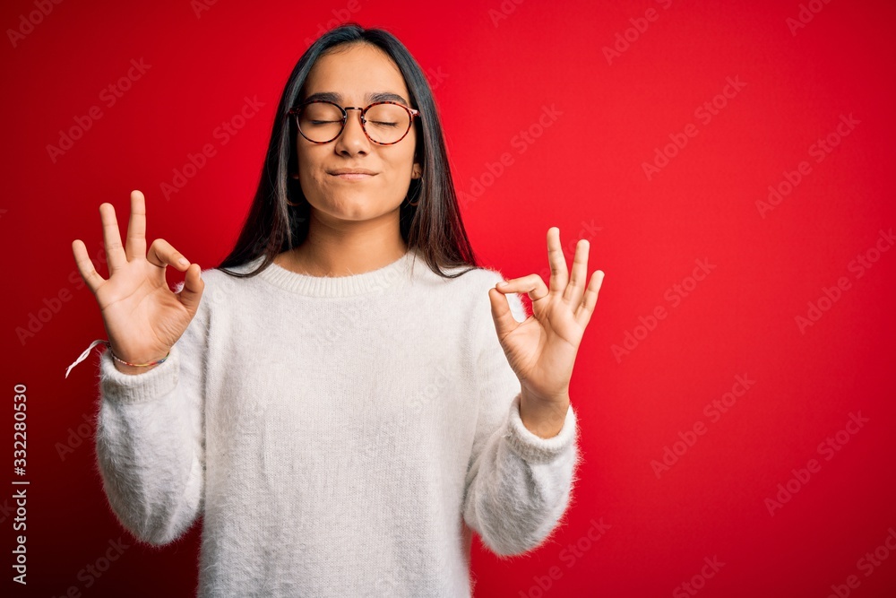 Young beautiful asian woman wearing casual sweater and glasses over red background relaxed and smiling with eyes closed doing meditation gesture with fingers. Yoga concept. Young beautiful asian woman wearing casual sweater and glasses over red background relaxed and smiling with eyes closed doing meditation gesture with fingers. Yoga concept.