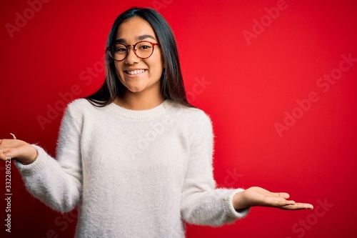 Young beautiful asian woman wearing casual sweater and glasses over red background smiling cheerful with open arms as friendly welcome, positive and confident greetings © Krakenimages.com