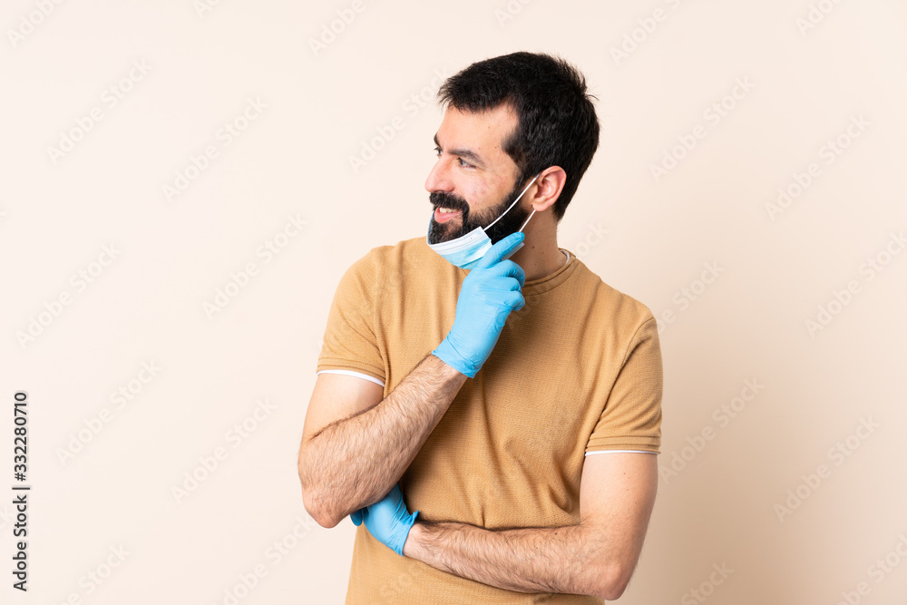 Caucasian man with beard protecting from the coronavirus with a mask and gloves over isolated background looking to the side and smiling