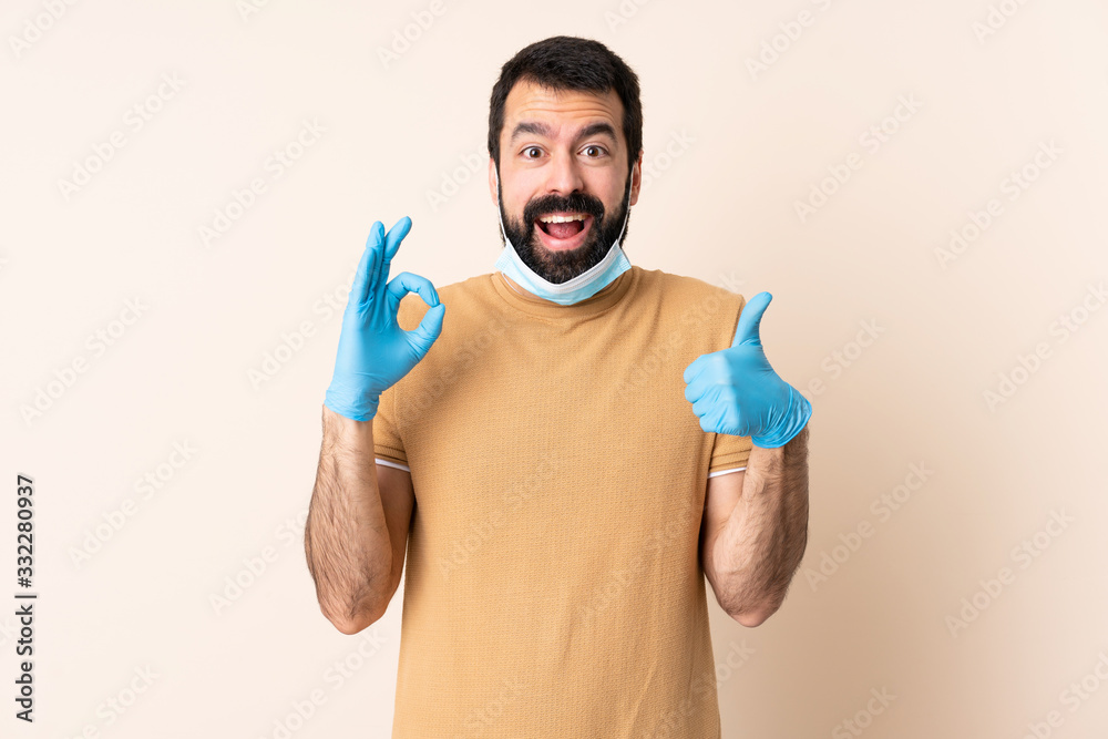 Caucasian man with beard protecting from the coronavirus with a mask and gloves over isolated background showing ok sign and thumb up gesture Caucasian man with beard protecting from the coronavirus with a mask and gloves over isolated background showing ok sign and thumb up gesture