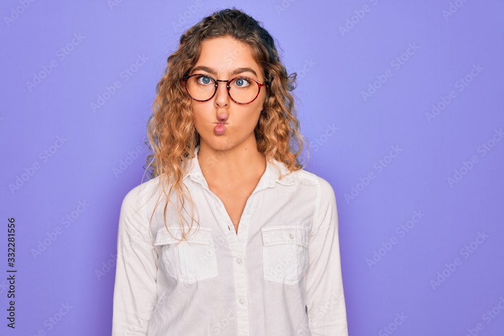Young beautiful woman with blue eyes wearing casual shirt and glasses over purple background making fish face with lips, crazy and comical gesture. Funny expression. Young beautiful woman with blue eyes wearing casual shirt and glasses over purple background making fish face with lips, crazy and comical gesture. Funny expression.