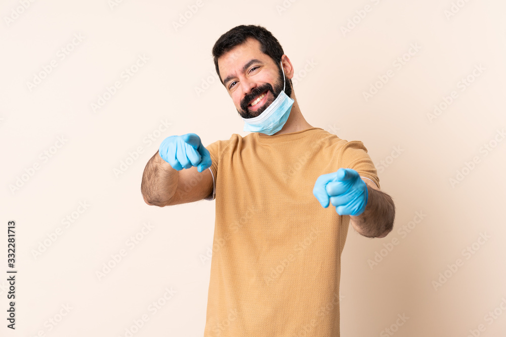 Caucasian man with beard protecting from the coronavirus with a mask and gloves over isolated background pointing front with happy expression Caucasian man with beard protecting from the coronavirus with a mask and gloves over isolated background pointing front with happy expression