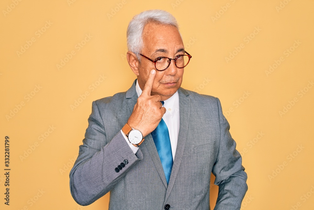 Middle age senior grey-haired handsome business man wearing glasses over yellow background Pointing to the eye watching you gesture, suspicious expression Middle age senior grey-haired handsome business man wearing glasses over yellow background Pointing to the eye watching you gesture, suspicious expression