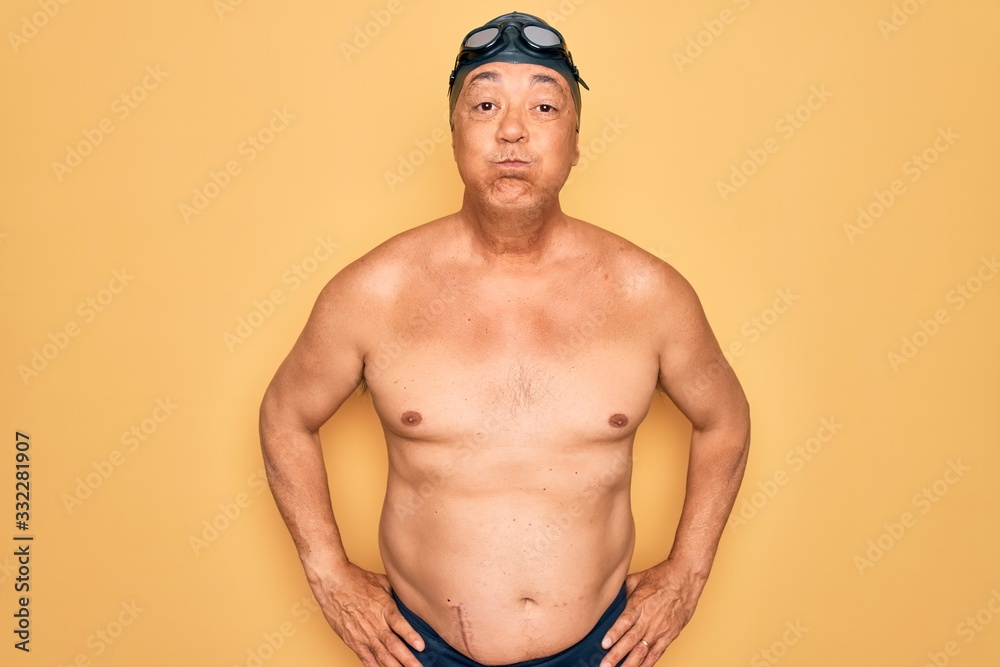 Middle age senior grey-haired swimmer man wearing swimsuit, cap and goggles puffing cheeks with funny face. Mouth inflated with air, crazy expression.