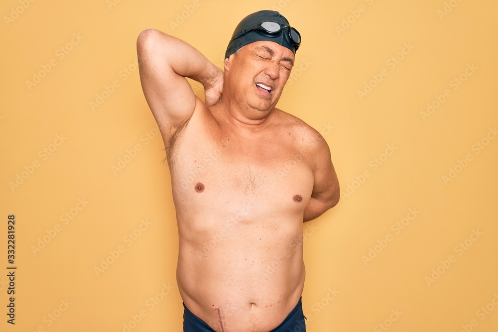 Middle age senior grey-haired swimmer man wearing swimsuit, cap and goggles Suffering of neck ache injury, touching neck with hand, muscular pain
