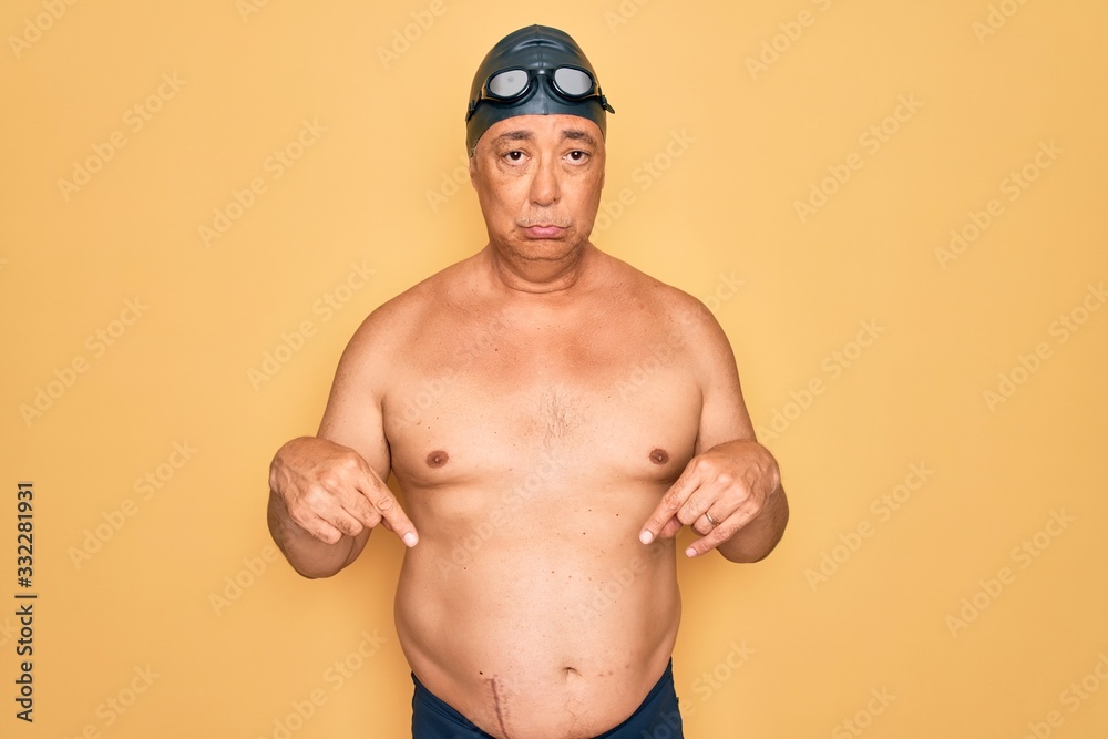 Middle age senior grey-haired swimmer man wearing swimsuit, cap and goggles Pointing down looking sad and upset, indicating direction with fingers, unhappy and depressed.
