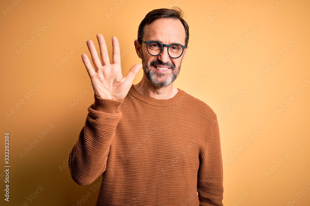 Middle age hoary man wearing brown sweater and glasses over isolated yellow background showing and pointing up with fingers number five while smiling confident and happy. Middle age hoary man wearing brown sweater and glasses over isolated yellow background showing and pointing up with fingers number five while smiling confident and happy.