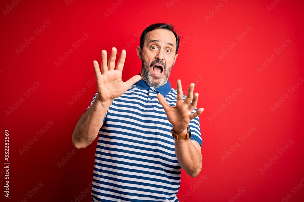 Naklejka premium Middle age hoary man wearing casual striped polo standing over isolated red background afraid and terrified with fear expression stop gesture with hands, shouting in shock. Panic concept.