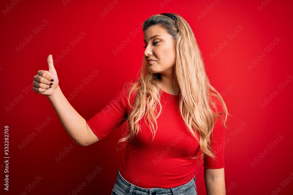 Young beautiful blonde woman wearing casual t-shirt standing over isolated red background Looking proud, smiling doing thumbs up gesture to the side Young beautiful blonde woman wearing casual t-shirt standing over isolated red background Looking proud, smiling doing thumbs up gesture to the side