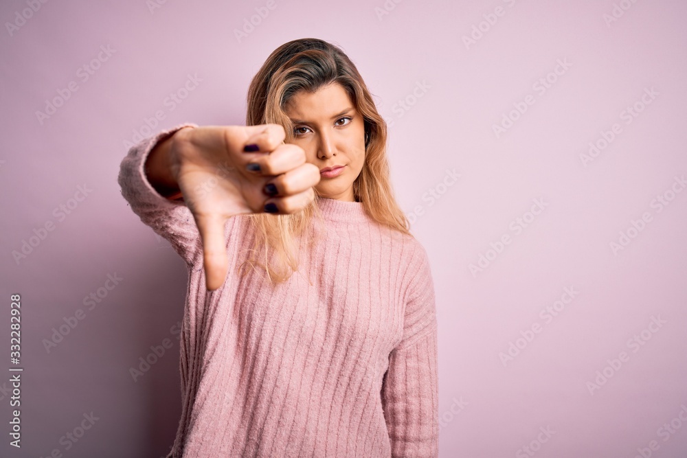 Young beautiful blonde woman wearing casual pink sweater over isolated background looking unhappy and angry showing rejection and negative with thumbs down gesture. Bad expression. Young beautiful blonde woman wearing casual pink sweater over isolated background looking unhappy and angry showing rejection and negative with thumbs down gesture. Bad expression.