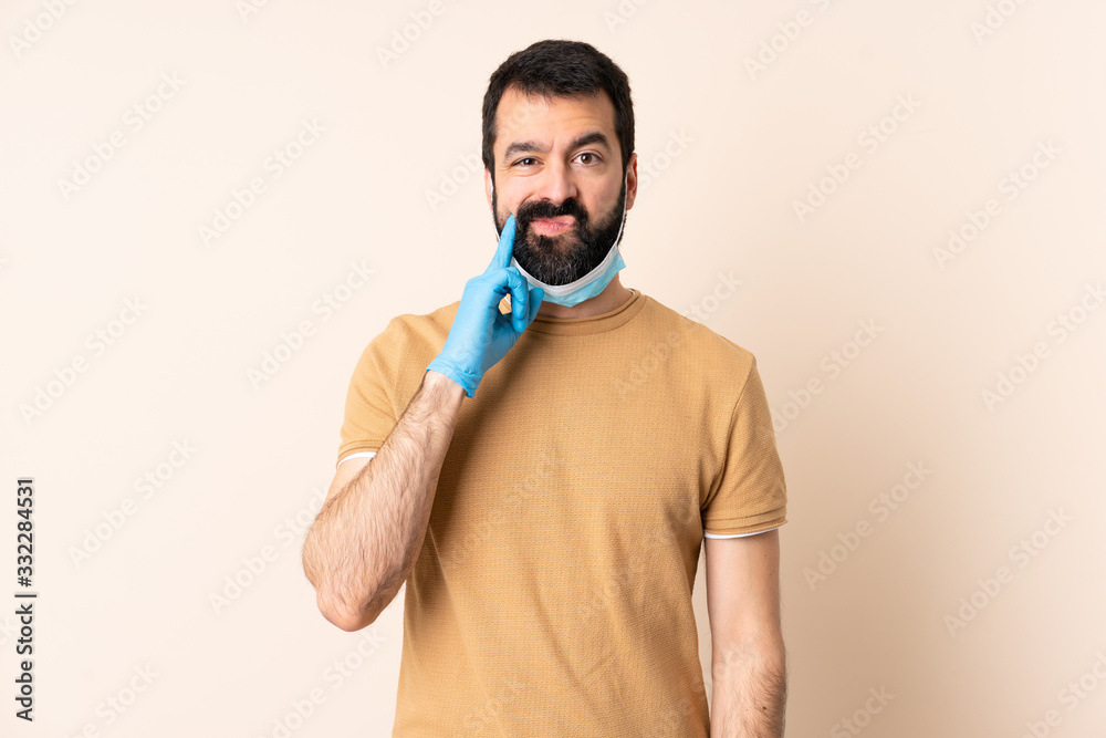 Caucasian man with beard protecting from the coronavirus with a mask and gloves over isolated background and thinking