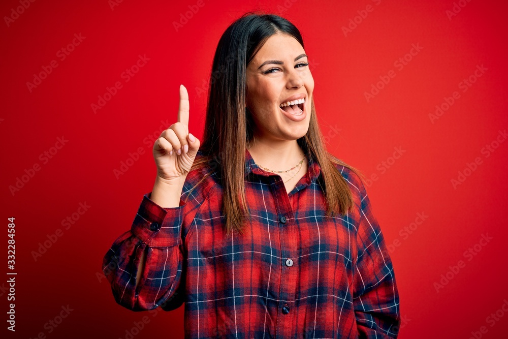 Young beautiful woman wearing casual shirt over red background pointing finger up with successful idea. Exited and happy. Number one. Young beautiful woman wearing casual shirt over red background pointing finger up with successful idea. Exited and happy. Number one.