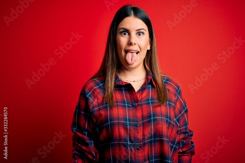 Young beautiful woman wearing casual shirt over red background sticking tongue out happy with funny expression. Emotion concept. © Krakenimages.com