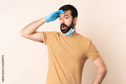 Caucasian man with beard protecting from the coronavirus with a mask and gloves over isolated background doing surprise gesture while looking to the side